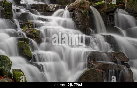 Long exposure of a waterfall with blurred water, wet rocks and green moss at The Cascade Waterfall at Virginia Water in Windsor Great Park, England Stock Photo