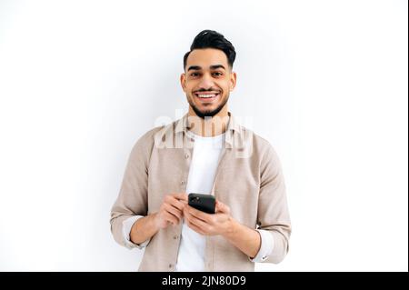 Handsome positive Arabian or Indian modern guy, holding smartphone in his hands, messaging, looking excitedly at the camera, standing over isolated white background, smiling Stock Photo