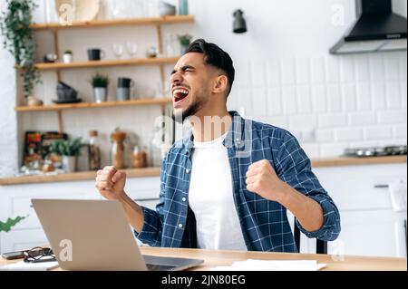 Cheerful emotional indian or arabian young man, trader, student, freelancer, sitting at table with laptop in kitchen, happy with good news, win, big profit, gesturing with fists, smiling, screaming Stock Photo