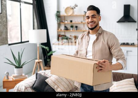 Happy confident indian or arabian guy, stand at home in living room, holding a large cardboard box, received a long-awaited parcel from the online store, preparing to unpack, looks at camera, smiles Stock Photo