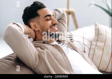 Chilling and relaxing at home. Handsome happy calm indian or arabian guy in casual clothes resting in the living room on the sofa, holds his hands behind his head with closed eyes and smiles Stock Photo