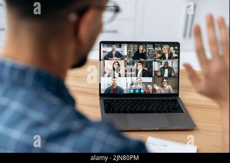Online video communication concept. Over shoulder view at a laptop screen with different people, employees, business partners, guy greets colleagues, online briefing, brainstorming, group conversation Stock Photo
