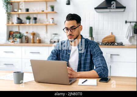 Serious focused arabian or indian guy with glasses, in stylish casual clothes, student, freelancer, company employee working remotely, sitting in the kitchen with a laptop, working on a project Stock Photo