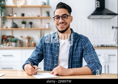Happy positive Arabian or Indian modern guy, clever student or freelancer, with glasses, listening to an online webinar, lecture, writing down information, looking at the camera, smiling friendly Stock Photo