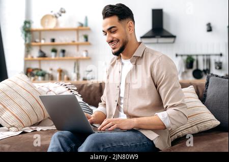 Modern successful smart indian or arabian freelancer, IT specialist, sits on a comfortable sofa in the living room, uses a laptop, works online, browsing the internet, social networks, smiles happily Stock Photo