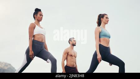 Yoga is all about being in the present moment. a group of young people standing and doing yoga while on the beach during an overcast day. Stock Photo