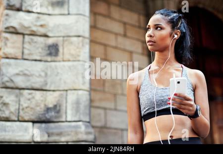 She moves to the beat of her own drum. an attractive young woman listening to music and using her cellphone while exercising outdoors in the city. Stock Photo