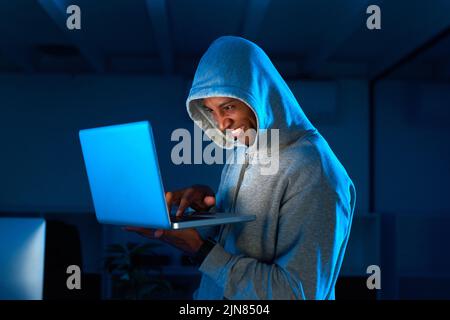 Firewall breached. a young male hacker smiling while using a laptop in the dark. Stock Photo