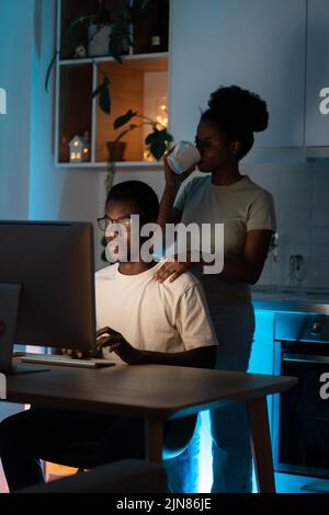 Black woman drinks coffee putting hand on shoulder of man in glasses sitting at table with computer Stock Photo