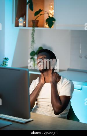 Tired Black man in white t-shirt kneads neck with hands sitting at table near computer in kitchen Stock Photo