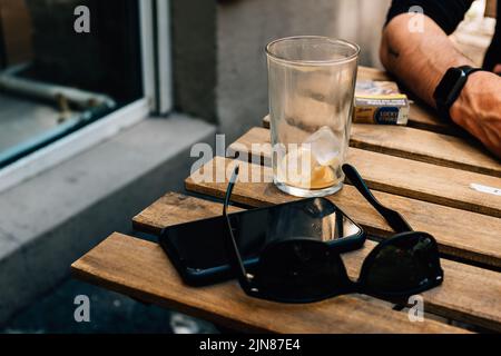 A glass with ice cubes, a smartphone, and shades are on a table beside a man Stock Photo