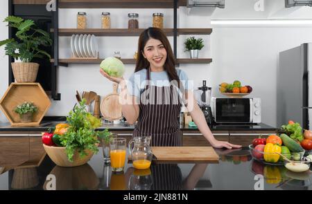 Young asian housewife dressed in an apron, hold fresh lettuce with one hand. The kitchen counter full of various kinds of vegetables. Stock Photo