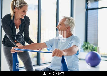 Youre doing a good job. a female physiotherapist helping a senior man stretch with a stretching band in a fitness centre. Stock Photo