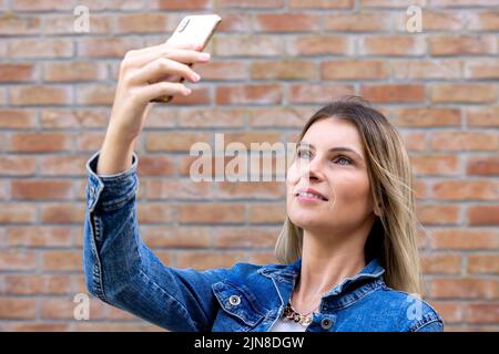 Photo of cheerful cute charming nice beautiful attractive girlfriend blonde, pale skinned taking selfie with her telephone held in her hands wearing jeans jacket isolated over red color brick background. High quality photo Stock Photo