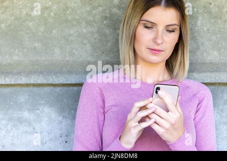 Full body photo of a young blonde caucasian sporty woman wearing pink sports apparel uses mobile phone in fitness center. female playing game on smartphone in gym. sporty girl social networking with smartphone in health club after working out. High quality photo Stock Photo