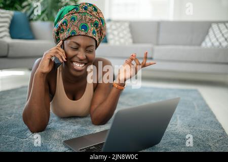 Beautiful African woman at home, smiling and laughing, using mobile smart phone, wearing traditional headscarf, surprised and happy Stock Photo