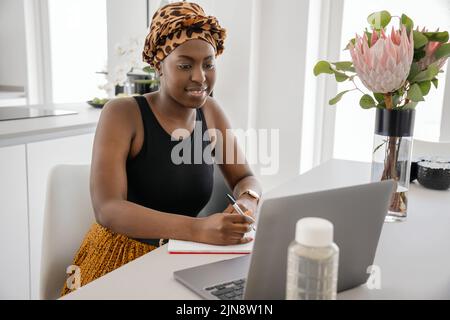 African woman studying from home, looking at laptop, writing down notes. Laptop and smartphone on home office table Stock Photo