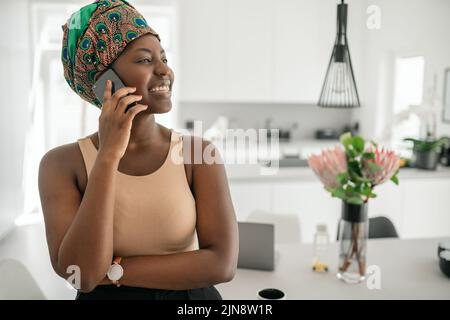 Beautiful African woman at home, smiling, using mobile smart phone, wearing traditional headscarf and looking into distance with copy space Stock Photo