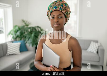 Portrait of beautiful African woman standing in modern home with laptop in hand looking into camera, wearing a traditional head tie scarf Stock Photo
