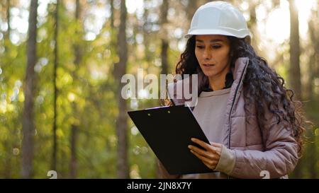 Millennial female technician ecologist looking up at treetops, Young indian woman in hardhat with clipboard taking measures checking trees. Forestry Stock Photo
