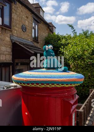 Red Royal Mail post box topped with a knitted frog, or yarn bombing, in the village of Hackleton, Northamptonshire, UK Stock Photo