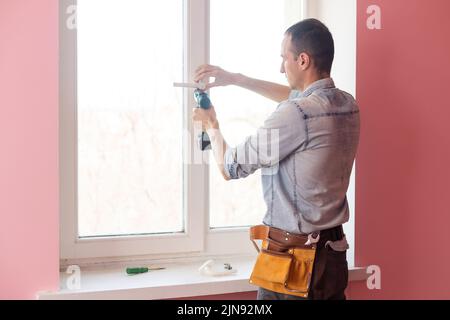 Handyman using a cordless screwdriver to install a window handle. Qualified worker services, home repair and renovation. Stock Photo