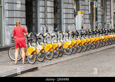 Lady in a red dress taking possession of a shared bicycle in a street in Brussels Stock Photo