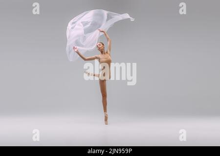 Portrait of young beautiful ballerina dancing with transparent fabric isolated over grey studio background. Grace Stock Photo