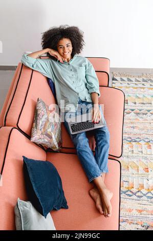 Top view at dark skin curly young woman web browsing or texting with friends online, using laptop sitting on the cozy sofa, female freelancer works remotely, looks at the camera and smiles Stock Photo
