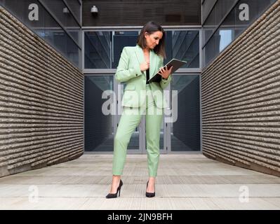 Full body of focused female entrepreneur in elegant suit and high heels reading documents while standing near building on street Stock Photo