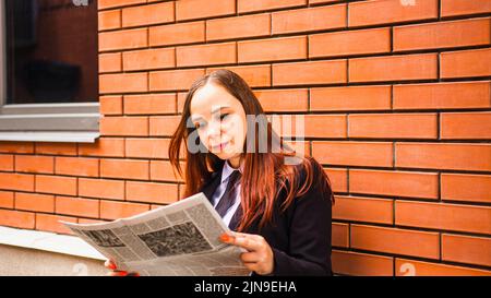 Young woman reading newspaper on street. Lady with long hair in casual clothes standing on street and reading newspaper. Stock Photo