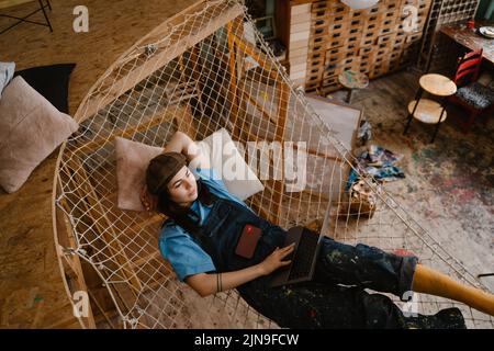 Young woman artist using laptop while resting in hammock at her studio indoors Stock Photo