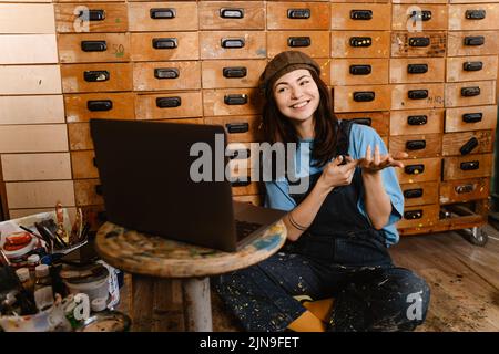 Young woman artist gesturing and using laptop while working in her studio indoors Stock Photo