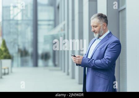 Serious and confident gray-haired man standing outside office building, businessman senior banker in business suit holding phone, successful investor using smartphone Stock Photo
