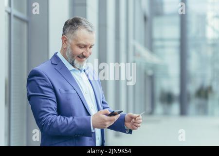 Senior gray-haired businessman outside office building, man in business suit uses mobile phone smartphone to buy in online store and order service, holds bank card in hands Stock Photo