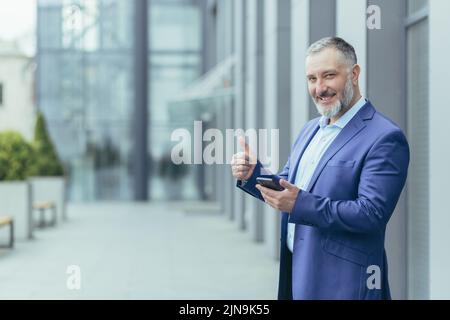 Portrait of successful senior gray haired banker, arab man looking at camera smiling and holding finger up, businessman outside office using smartphone Stock Photo