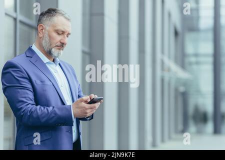 Serious and confident gray-haired man standing outside office building, businessman senior banker in business suit holding phone, successful investor using smartphone Stock Photo