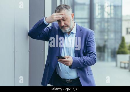 Senior gray-haired businessman in despair upset reading bad news on phone, man outside office building in business suit, bankrupt investor lost money, banker in depression Stock Photo