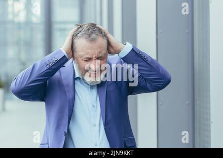 Senior gray-haired businessman outside office building depressed, man in despair and despondency, bankrupt investor lost money, holding hands on head Stock Photo
