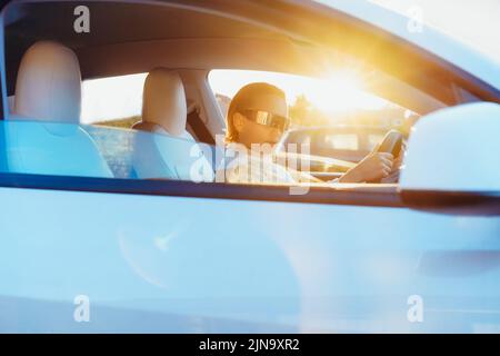 Portrait of fashion cyberpunk girl driving a white supercar in sunsert light. Woman with futuristic eyeglasses sits by the car steering wheel. Future Stock Photo