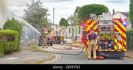 Essex Fire and Rescue Service firemen spraying water jet on house two fire brigade fire engines & firefighters in residential street scene England UK Stock Photo