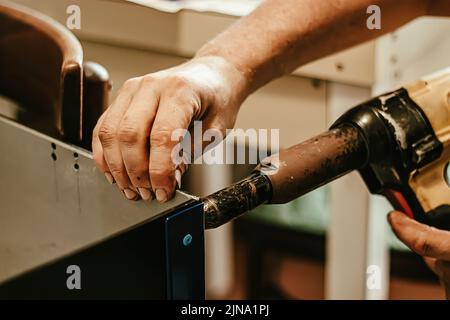 Male worker uses electric riveting gun. Hand holding  riveting machine and placing rivets. Working workshop of carpenter or mechanic. Stock Photo