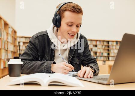 Smiling college student wearing wireless headphones while writing on exercise book in library. Guy with ginger hair doing homework. Stock Photo