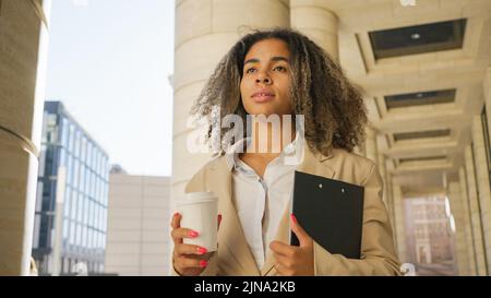 Serious black business woman in a white shirt and jacket goes to a meeting with a cup of coffee and a folder with documents against the backdrop of a modern business center Stock Photo