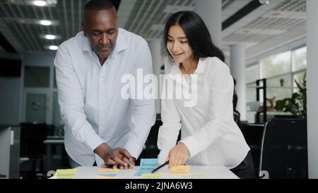 Two colleagues african and arab businesspeople co-workers writing tasks on sticky notes brainstorming organizing work together managing paperwork in Stock Photo