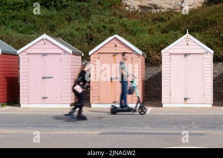 row of multicoloured pink wooden beach huts by the sea withskater and man on electric scooter  passing in front, in Bournemouth Dorset Uk Stock Photo