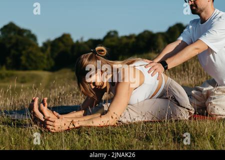 Man Helping Woman Doing Stretching Outdoors, Young Adult Couple Practicing Yoga Together Stock Photo