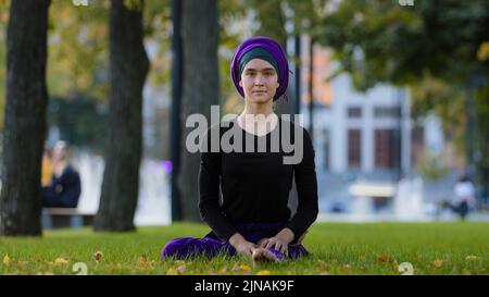 Young girl muslim woman in hijab sits on green grass in park takes leg by foot bends turns to side stretching doing asana twisting body core yoga Stock Photo