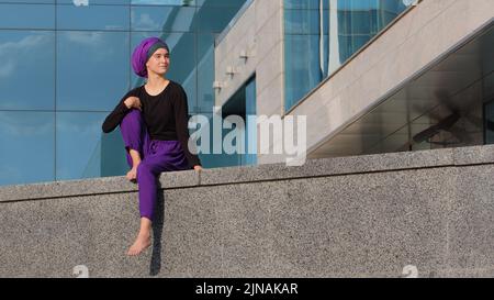 Calm young muslim girl buddist in hijab lonely lady female beautiful woman sits on building city skyscraper background looking around waiting dreaming Stock Photo