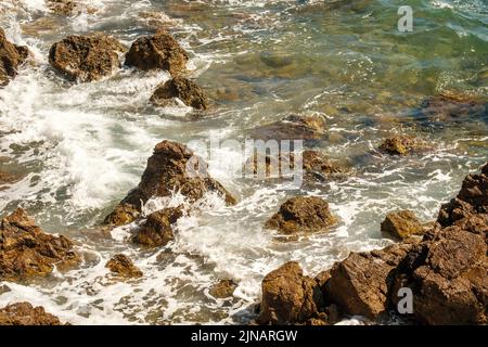 Small ocean waves crashing of rocks on shallow coastline at high tide at bright sunlight. Transparent waves create foams rolling on stones Stock Photo